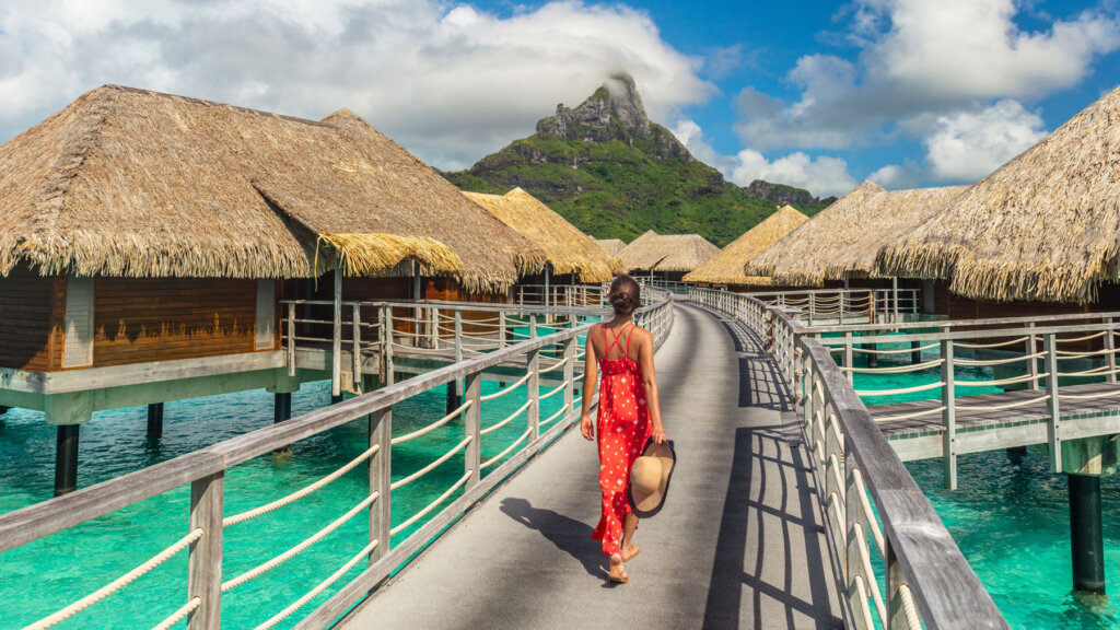 Luxury travel advisor client walking along overwater bungalows at a tropical resort with turquoise lagoon and mountain backdrop